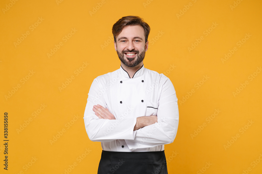 Smiling young bearded male chef cook or baker man in white uniform shirt posing isolated on yellow orange background studio portrait. Cooking food concept. Mock up copy space. Holding hands crossed. - obrazy, fototapety, plakaty 