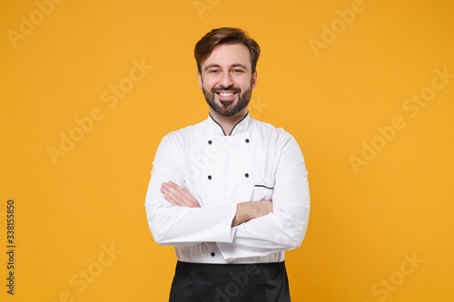 Smiling young bearded male chef cook or baker man in white uniform shirt posing isolated on yellow orange background studio portrait. Cooking food concept. Mock up copy space. Holding hands crossed.