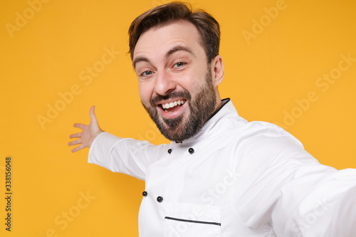 Close up of excited bearded male chef cook or baker man in white uniform isolated on yellow background. Cooking food concept. Mock up copy space. Doing selfie shot on mobile phone pointing hand aside.