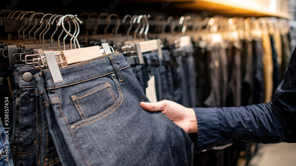 Male hand customer choosing black men jeans or denim pants (trousers)  hanging on rack in clothes shop. Fashion product collection in clothing  store for selling. Textile industry and business concept Photos