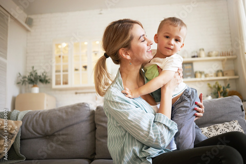 Indoor image of cute young female with ponytail holding tight her charming baby, sitting on sofa with him. Pretty mother and son bonding in living room, mom looking at child with love and tenderness