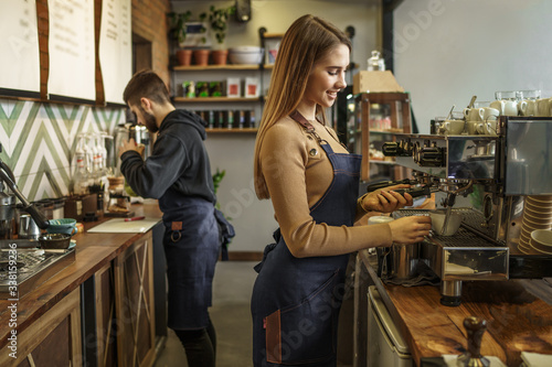 Professional female barista holding metal jug warming milk using the coffee machine. Positive smiling woman preparing coffee at machine