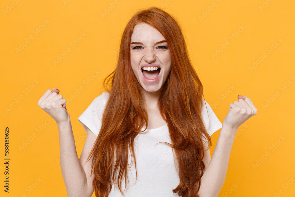 Overjoyed young redhead woman girl in white blank empty t-shirt posing isolated on yellow wall background studio portrait. People lifestyle concept. Mock up copy space. Doing winner gesture screaming. Overjoyed young redhead woman girl in white blank empty t-shirt posing isolated on yellow wall background studio portrait. People lifestyle concept. Mock up copy space. Doing winner gesture screaming.