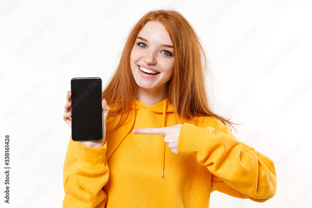 Smiling young redhead woman girl in yellow hoodie posing isolated on white wall background. People lifestyle concept. Mock up copy space. Pointing index finger on mobile phone with blank empty screen. Smiling young redhead woman girl in yellow hoodie posing isolated on white wall background. People lifestyle concept. Mock up copy space. Pointing index finger on mobile phone with blank empty screen.