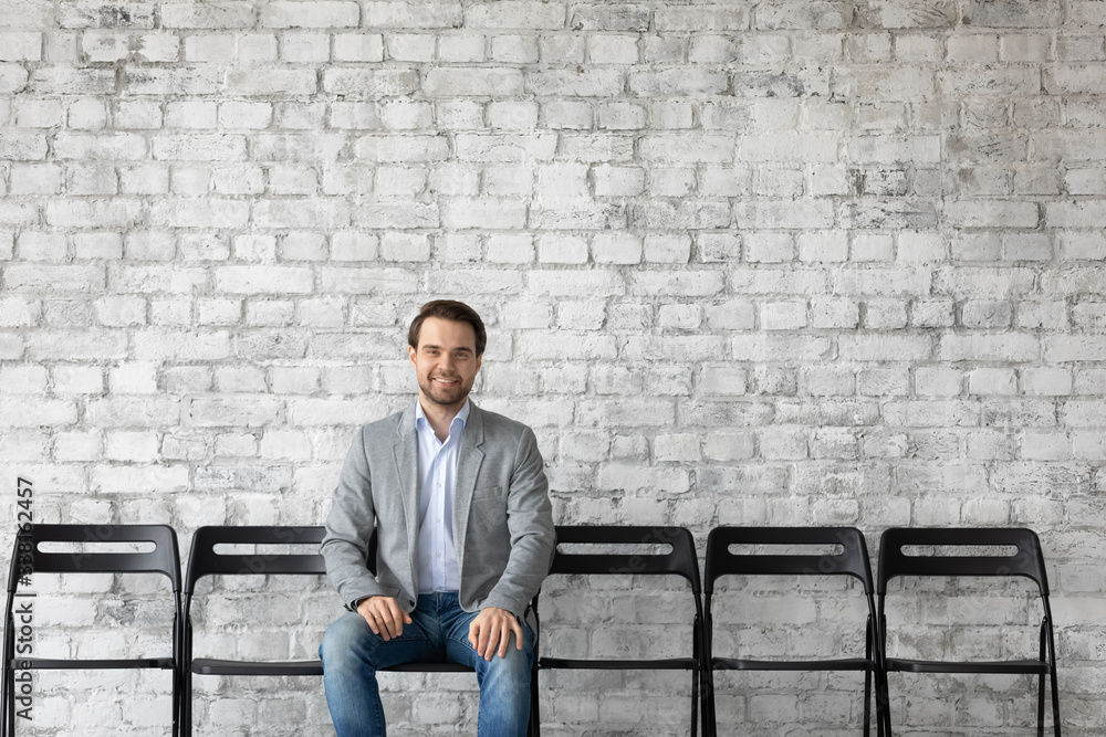 Portrait of smiling Caucasian male job candidate sit on chair in hallway wait for interview in office, happy man work applicant apply at vacant position at workplace, hiring, employment concept Portrait of smiling Caucasian male job candidate sit on chair in hallway wait for interview in office, happy man work applicant apply at vacant position at workplace, hiring, employment concept