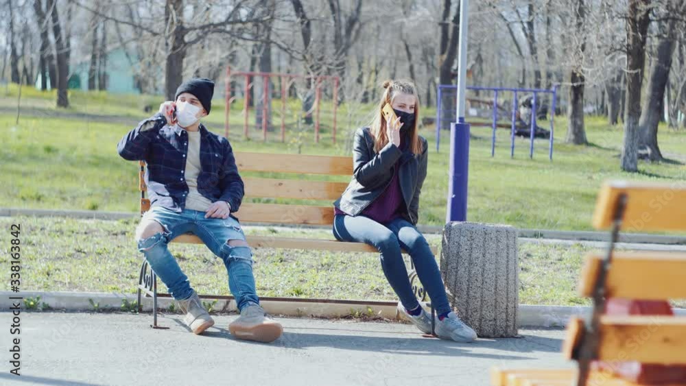 a young couple sits on a bench in medical masks in the distance and talking on the phone with each other a young couple sits on a bench in medical masks in the distance and talking on the phone with each other
