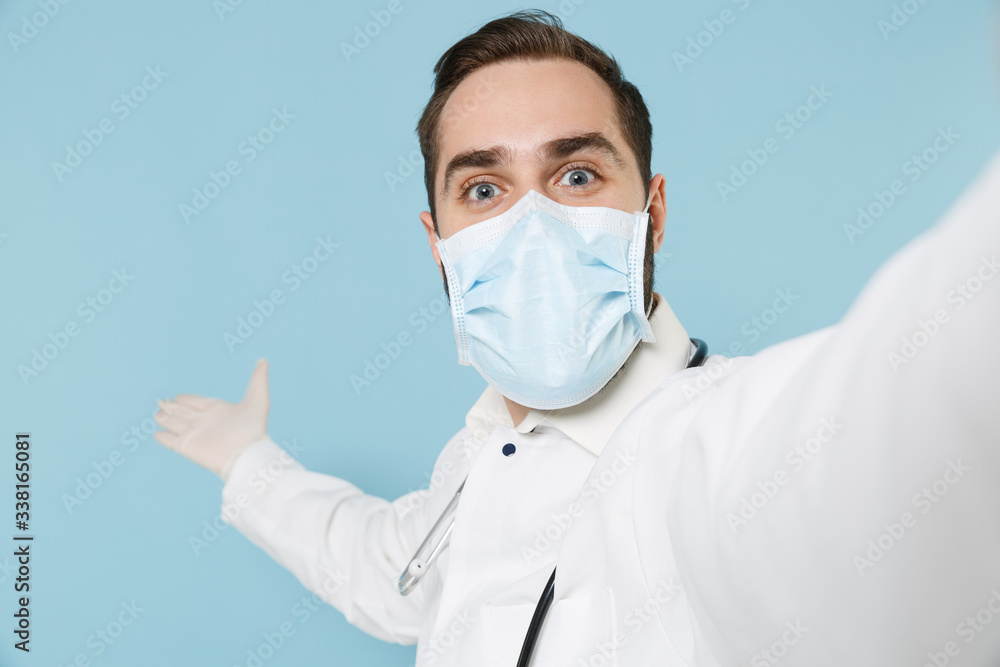 Close up of doctor man in medical gown face mask gloves isolated on blue background. Epidemic pandemic coronavirus 2019-ncov sars covid-19 flu virus. Doing selfie on mobile phone pointing hand aside. Close up of doctor man in medical gown face mask gloves isolated on blue background. Epidemic pandemic coronavirus 2019-ncov sars covid-19 flu virus. Doing selfie on mobile phone pointing hand aside.