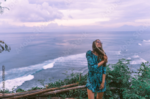 A beautiful girl stands on a cliff overlooking the waves of the blue ocean. Tropical paradise and exotic nature on the island of Bali. Model with different emotions of happiness and joy.