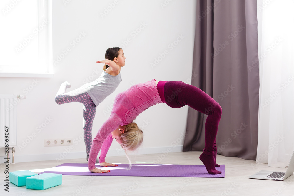 Fototapeta premium Mother and daughter practicing online yoga lesson at home at quarantine isolation period during coronavirus pandemic. Family doing sport together online from home. Healthy lifestyle