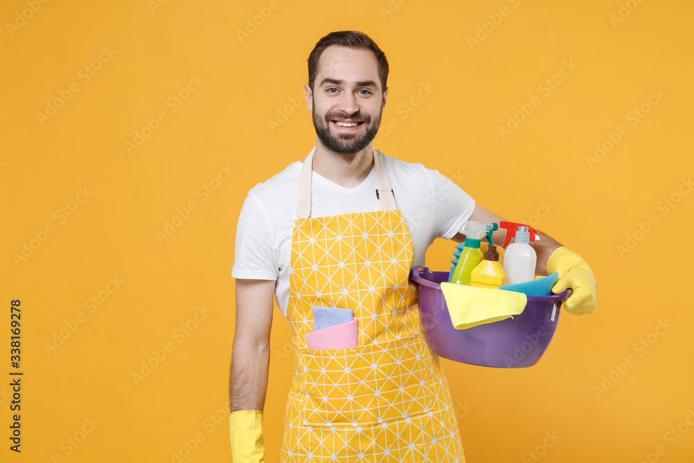 Smiling young man househusband in apron rubber gloves hold basin with detergent bottles washing cleansers doing housework isolated on yellow background studio. Housekeeping concept. Looking camera. Smiling young man househusband in apron rubber gloves hold basin with detergent bottles washing cleansers doing housework isolated on yellow background studio. Housekeeping concept. Looking camera.