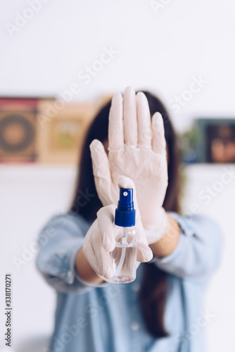 Coronavirus concept. Girl in her home wear gloves and show as an antiseptic spray, says stop with her hand to coronavirus. Global call to stay home.