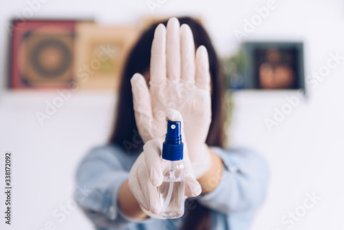 Fototapeta Naklejka Na Ścianę i Meble -  Coronavirus concept. Girl in her home wear gloves and show as an antiseptic spray, says stop with her hand to coronavirus. Global call to stay home.