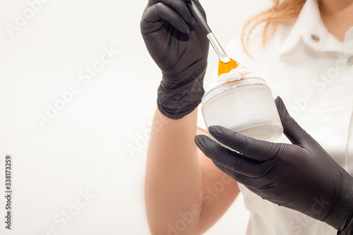 A jar of cream, white clay or face mask in the hands of a cosmetologist, in black latex gloves, cream on a special brush for applying to the face. The concept of skin care.