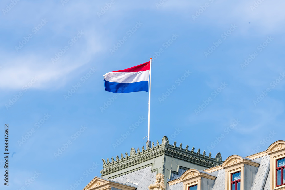 Flag of the Kingdom of the Netherlands in red, white and blue strips of fabric fastened at a flagpole on a building and blowing in the wind.