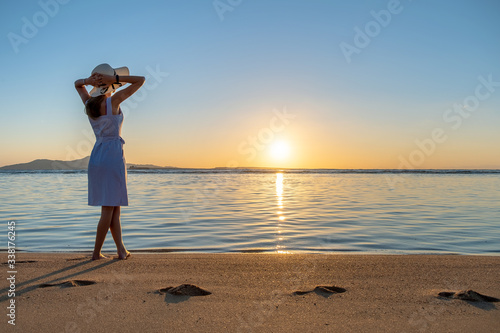 Young woman in straw hat and a dress standing alone on empty sand beach at sunset sea shore. Lonely girl looking at horizon over calm ocean surface on vacation trip.