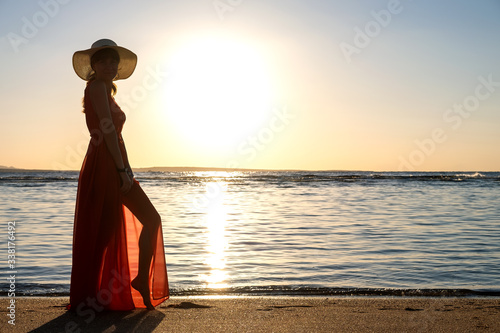 Young woman wearing long red dress and straw hat standing on sand beach at sea shore enjoying view of rising sun in early summer morning.