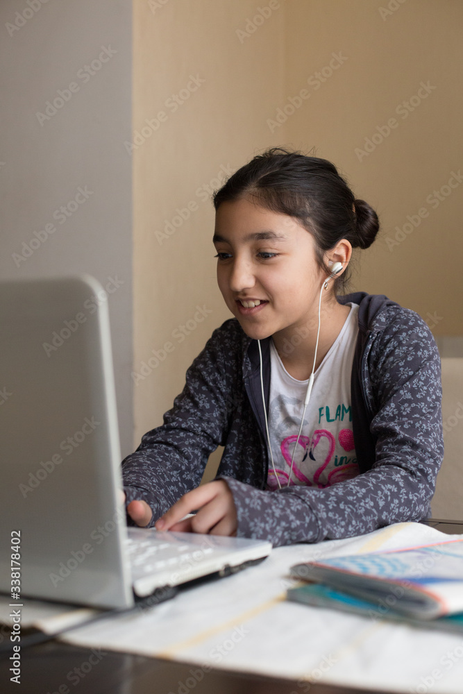Young girl sitting at the dining table with laptop at home schooling, online virtual classroom video conference, distant education. Active participation at the lesson with earphones, doing homework. Young girl sitting at the dining table with laptop at home schooling, online virtual classroom video conference, distant education. Active participation at the lesson with earphones, doing homework.