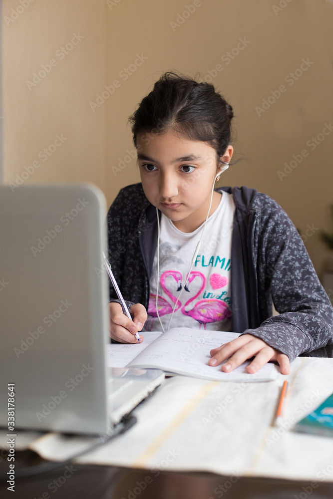 Young girl sitting at the dining table with laptop at home schooling, online virtual classroom video conference, distant education. Active participation at the lesson with earphones, doing homework. Young girl sitting at the dining table with laptop at home schooling, online virtual classroom video conference, distant education. Active participation at the lesson with earphones, doing homework.