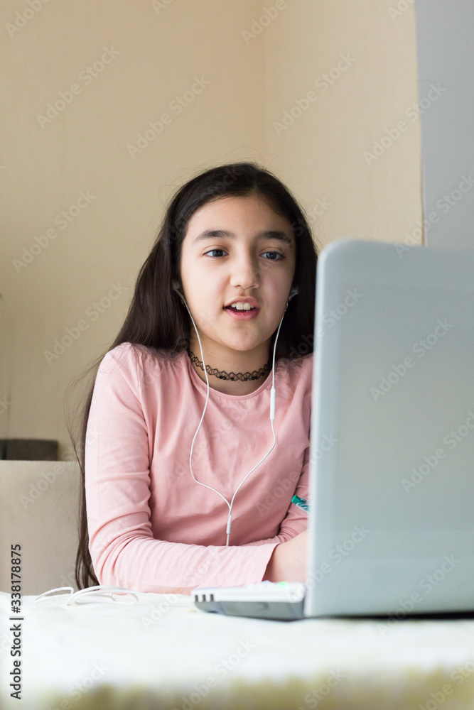 Young girl sitting at the dining table with laptop at home schooling, online virtual classroom video conference, distant education. Active participation at the lesson with earphones, doing homework. Young girl sitting at the dining table with laptop at home schooling, online virtual classroom video conference, distant education. Active participation at the lesson with earphones, doing homework.