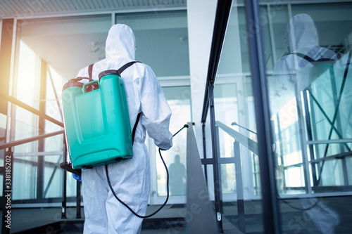Unrecognizable person in white protection suit disinfecting public areas to stop spreading highly contagious coronavirus. Man with tank reservoir on his back spraying disinfectant to kill COVID-19.