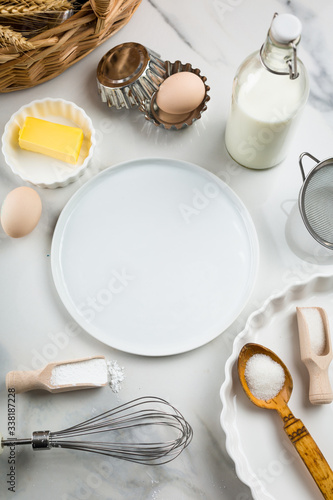 Ingredients for sweet baking on white marble background, eggs, milk jar, sugar, flour, butter, baking powder. Cookies or cake baking praparation. White round mock up for text in the center, copy space