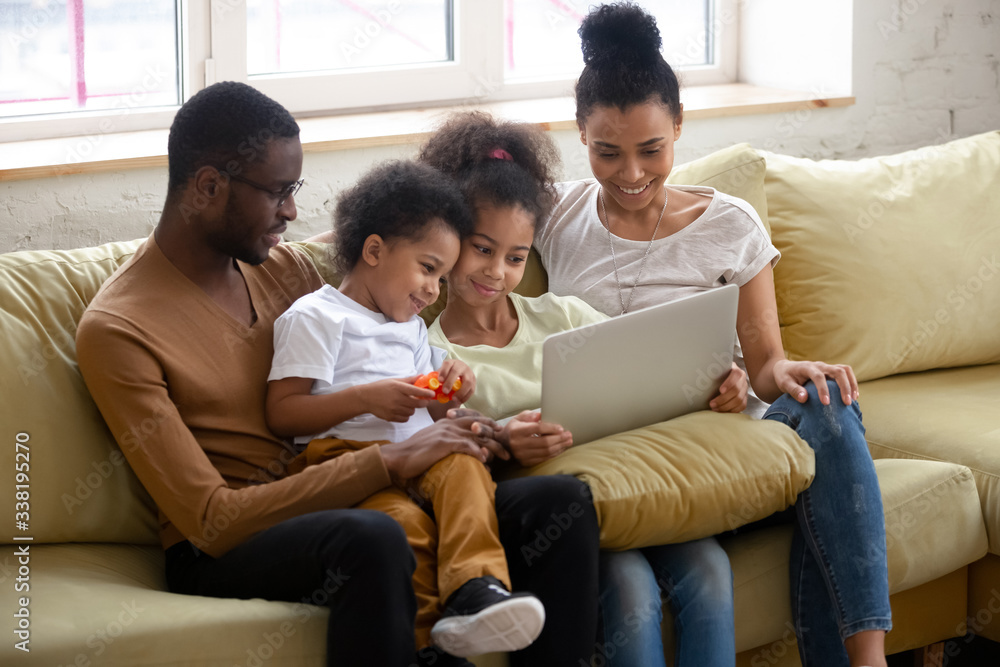 African american happy family with two children having good time using laptop. Young diverse smiling husband and wife with cute daughter and son sitting on couch at home looking at computer.