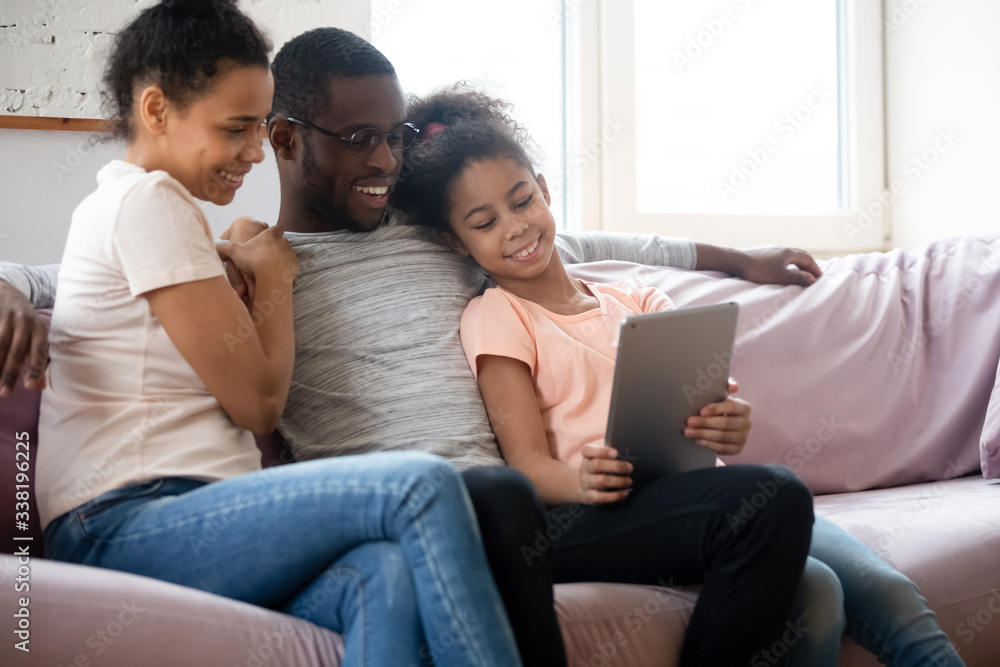 Happy african american couple looking at tablet screen with daughter. Diverse smiling family of mother, father and girl sitting on couch using mobile device and gadget together.