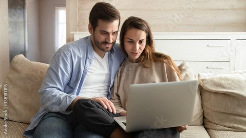 Young affectionate man cuddling mixed race girlfriend, watching funny movie in comfortable living room. Happy millennial married couple relaxing on cozy sofa, looking at computer monitor at home.