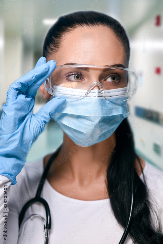Girl in protective glasses, medical mask and blue gloves on blured hospital background . close-up portrait of a woman in a transparent mask. way to protect against coronavirus. Covid-2019, Pandemic 20