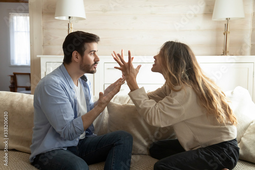 Emotional annoyed stressed couple sitting on couch, arguing at home. Angry irritated nervous woman man shouting at each other, figuring out relations, feeling outraged, relationship problems concept. photo