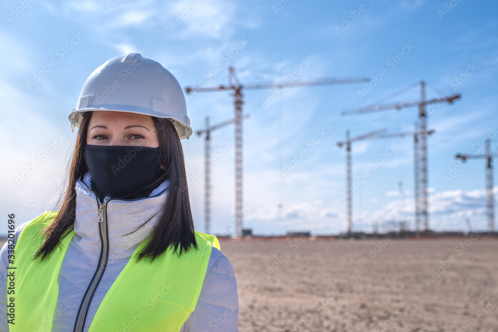 a female construction worker in a black mask, white helmet and yellow vest on the background of a construction site a female construction worker in a black mask, white helmet and yellow vest on the background of a construction site