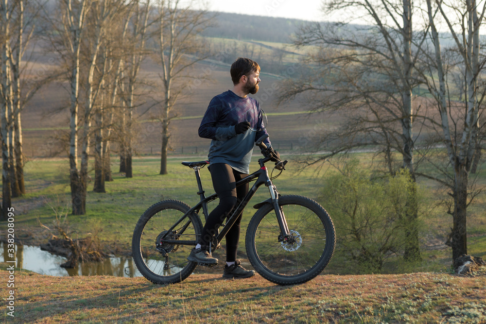 Cyclist in shorts and jersey on a modern carbon hardtail bike with an air suspension fork standing on a cliff against the background of fresh green spring forest Cyclist in shorts and jersey on a modern carbon hardtail bike with an air suspension fork standing on a cliff against the background of fresh green spring forest