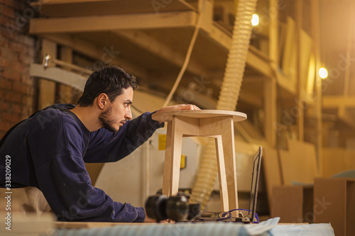 portrait of young cerpenter checking handmade wooden chair after grinding in workshop. confident woodworker make furniture photo