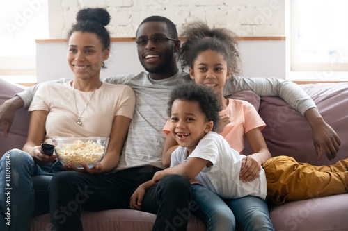Happy african american family with kids watching funny tv show or movie eating popcorn snack. Happy diverse dad holding remote controller, mom hugging cute siblings.