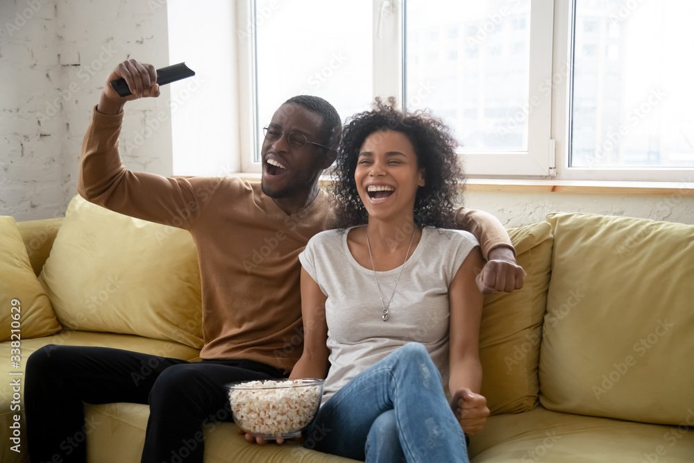 Young happy african american couple watching funny movie or tv show eating popcorn snack. Attractive smiling family of diverse woman and man sitting on sofa holding remote controller. Young happy african american couple watching funny movie or tv show eating popcorn snack. Attractive smiling family of diverse woman and man sitting on sofa holding remote controller.