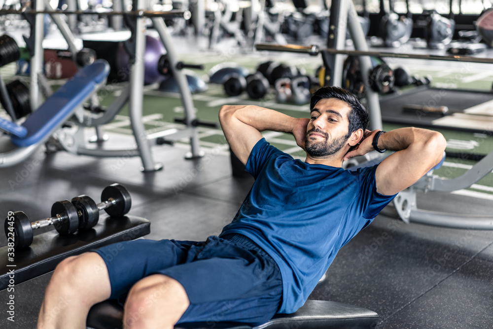 Naklejka premium Young fit handsome smiling Caucasian man exercising by doing a sit-up on bench in gym or fitness club. Male athlete has good body shape with six pack. Sport recreation and fitness for health concept.