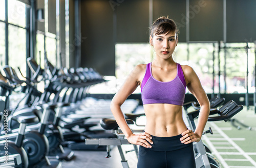 Portrait of young pretty Asian girl with six packs in purple color sportswear standing and crossing arms in gym or fitness club. Behind her is exercise machines and equipment. Fitness & Gym concept.