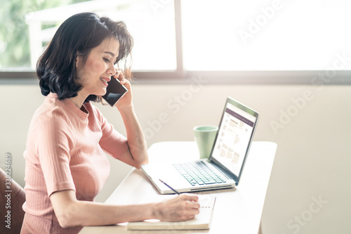Asian young business woman sit and write note in a notebook while talk on phone and open files in laptop. During Coronavirus crisis Covid19 outbreak girl work from home for prevent contract a disease.