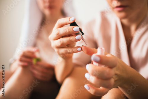 Two young women in towels and pajamas have a fun spa party together in home. They are sitting on the bed and making themselves a new Manicure