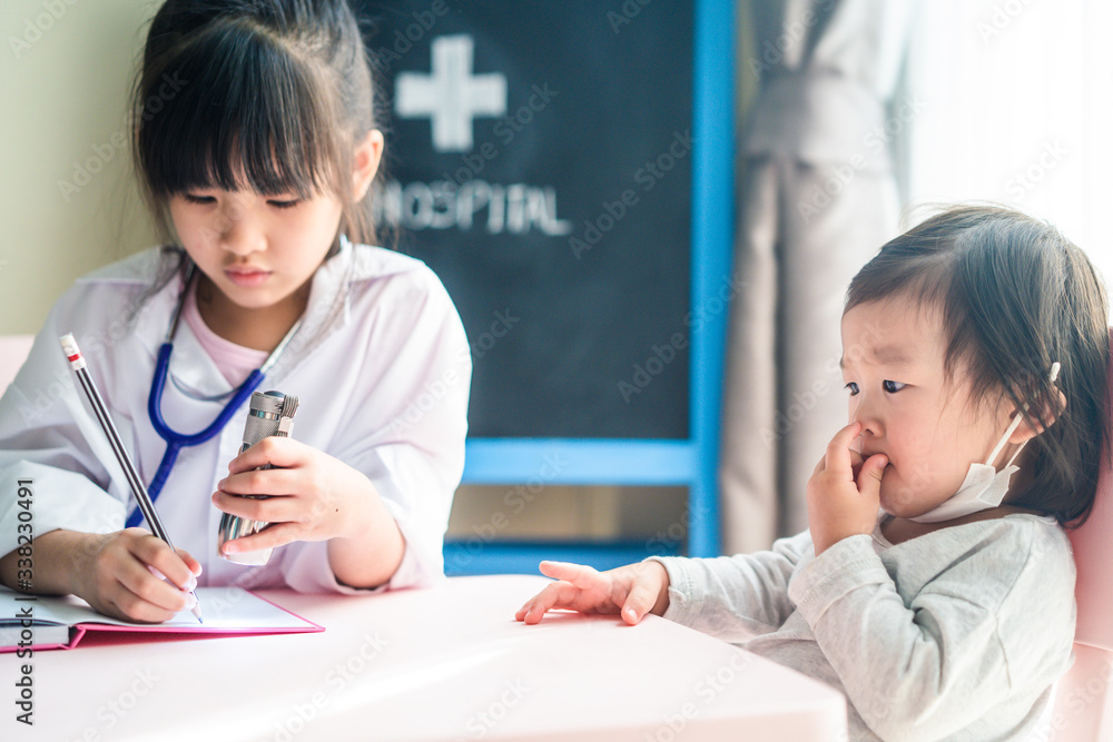 Two Asian cute girls pretend to be doctor and patient. Young doctor write diagnosis disease in notebook, give some advice to little sick baby who taking some medicine and vitamin in examination room. Two Asian cute girls pretend to be doctor and patient. Young doctor write diagnosis disease in notebook, give some advice to little sick baby who taking some medicine and vitamin in examination room.