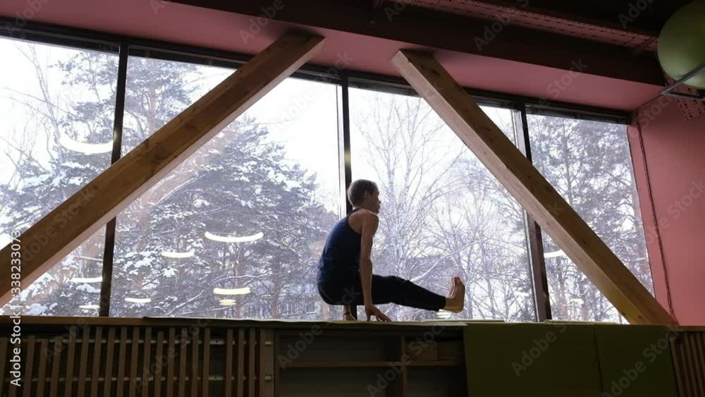 a nice man does yoga on the windowsill of a large window. Outside the window is a winter forest and daylight. Wide-angle shooting a nice man does yoga on the windowsill of a large window. Outside the window is a winter forest and daylight. Wide-angle shooting