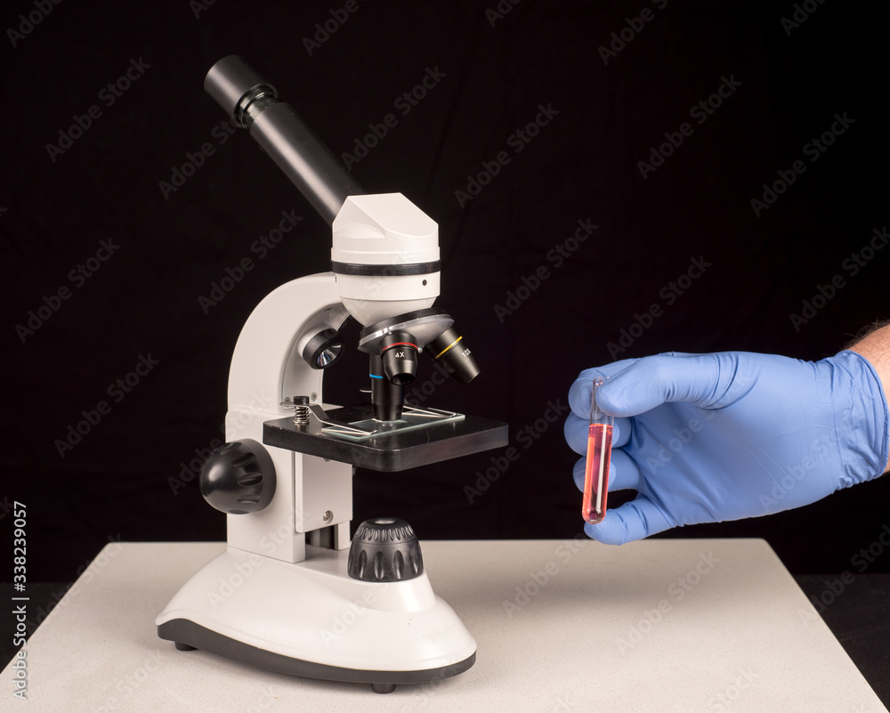 Microscope and hand with surgical glove holding vial with red solution. Black background with space for text Microscope and hand with surgical glove holding vial with red solution. Black background with space for text