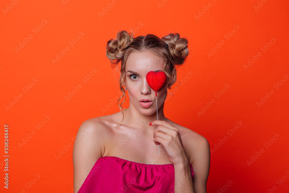 The girl in a red dress on a orange background in the studio. Blonde girl with two hair knots holding red heart on the stick, looking to the camera and smiling. The girl in a red dress on a orange background in the studio. Blonde girl with two hair knots holding red heart on the stick, looking to the camera and smiling.