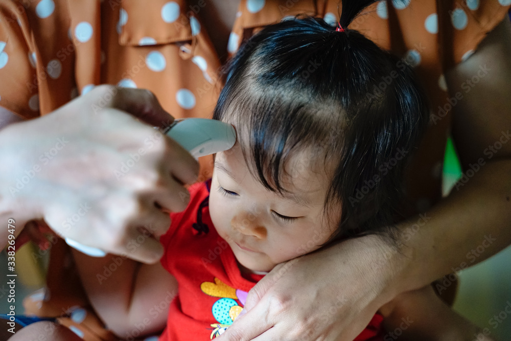 Mother measuring temperature cute baby girl at home . Mother takes temperature for toddler with Infrared Forehead Thermometer for virus symptomnm.Coronavirus screening concept. Mother measuring temperature cute baby girl at home . Mother takes temperature for toddler with Infrared Forehead Thermometer for virus symptomnm.Coronavirus screening concept.