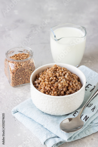 Tasty buckwheat porridge in a white ceramic bowl and a jug with milk, light grey concrete background. Idea for healthy gluten free breakfast.