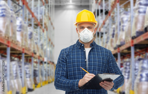 health care, work and pandemic concept - male worker in safety helmet wearing face protective mask or respirator with clipboard and pencil over warehouse background