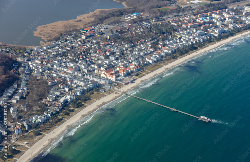 The baltic seaside resort of Binz on the German island of Rügen was photographed from the air. Below is the beach and the pier. A white excursion boat docks there. In the background the train station.