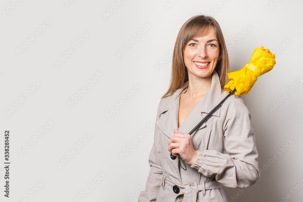Smiling cheerful young woman in a beige raincoat, holds a rolled-up yellow umbrella on her shoulder on a light background. Concept weather forecast, rain. Empty space for text Smiling cheerful young woman in a beige raincoat, holds a rolled-up yellow umbrella on her shoulder on a light background. Concept weather forecast, rain. Empty space for text