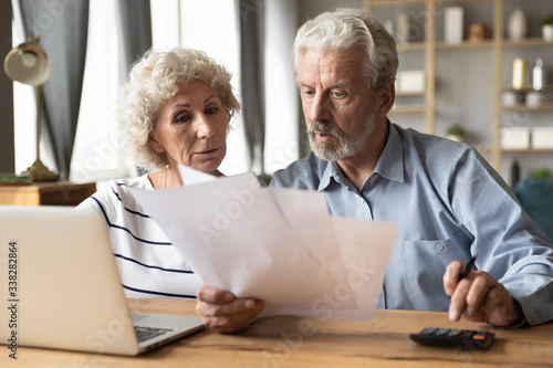 Serious older couple reading financial documents, planning budget or checking bills, mature man using calculator, counting taxes, senior husband and wife discussing bank debt or loan payment