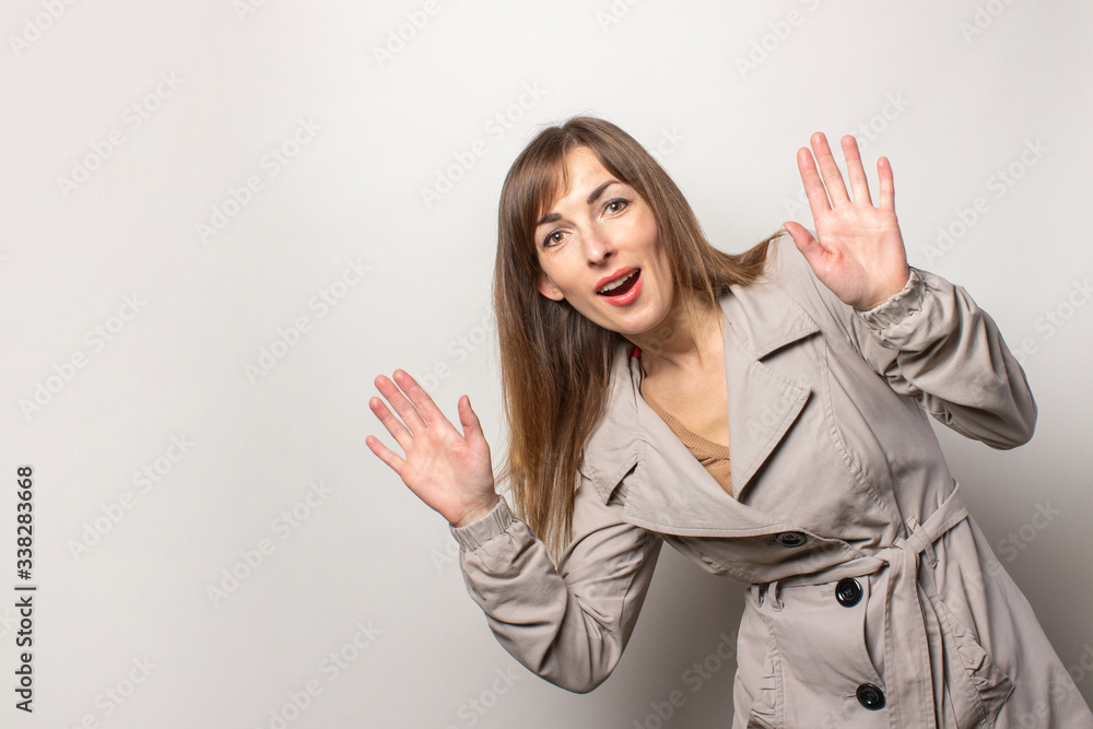 cheerful young women looks out from the side and holds her hands in front of her and looks at the camera on a light background. The model is wearing a beige cloak. Concept style, women's fashion cheerful young women looks out from the side and holds her hands in front of her and looks at the camera on a light background. The model is wearing a beige cloak. Concept style, women's fashion
